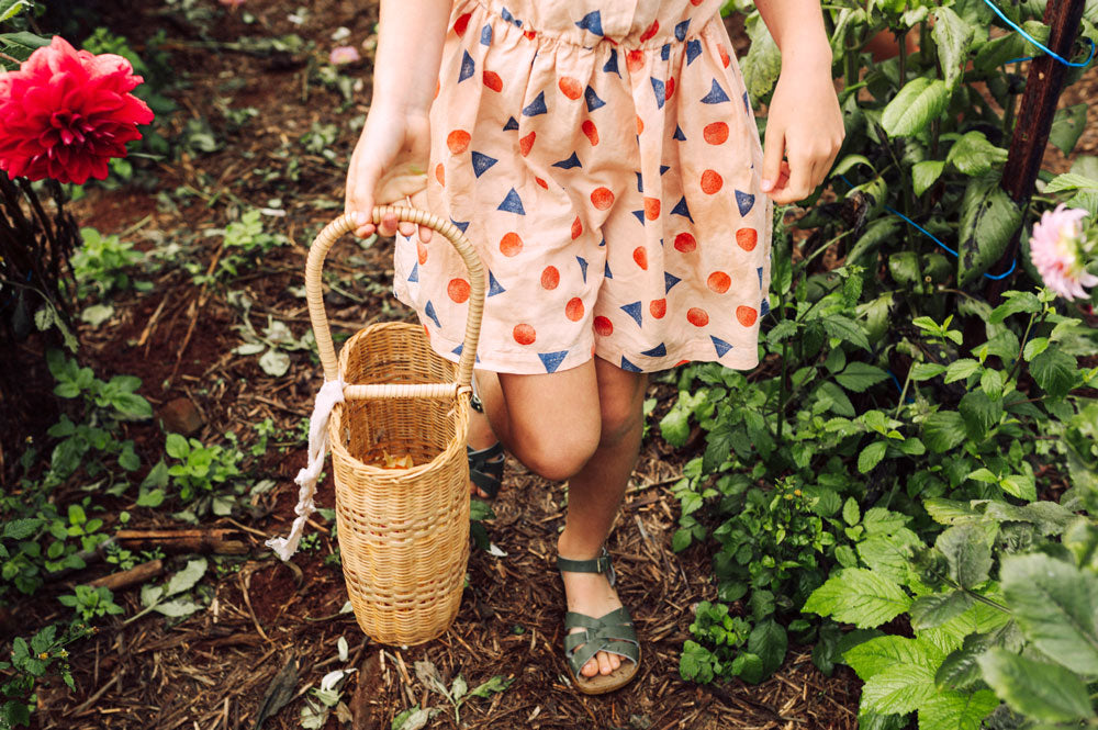 A girl wears green sandals and a pattern dress in the garden while holding a basket