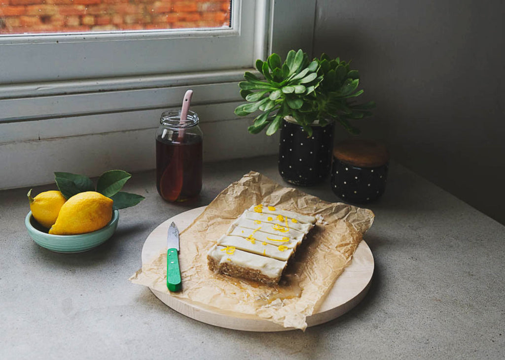 a cutting board with no-bake lemon and coconut slice on it beside a window, next to a bowl of lemons