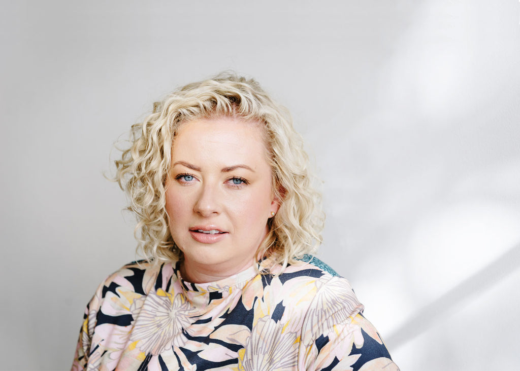 Portrait of Danielle Binks, a caucasian woman with blonde curly hair wearing a floral shirt.