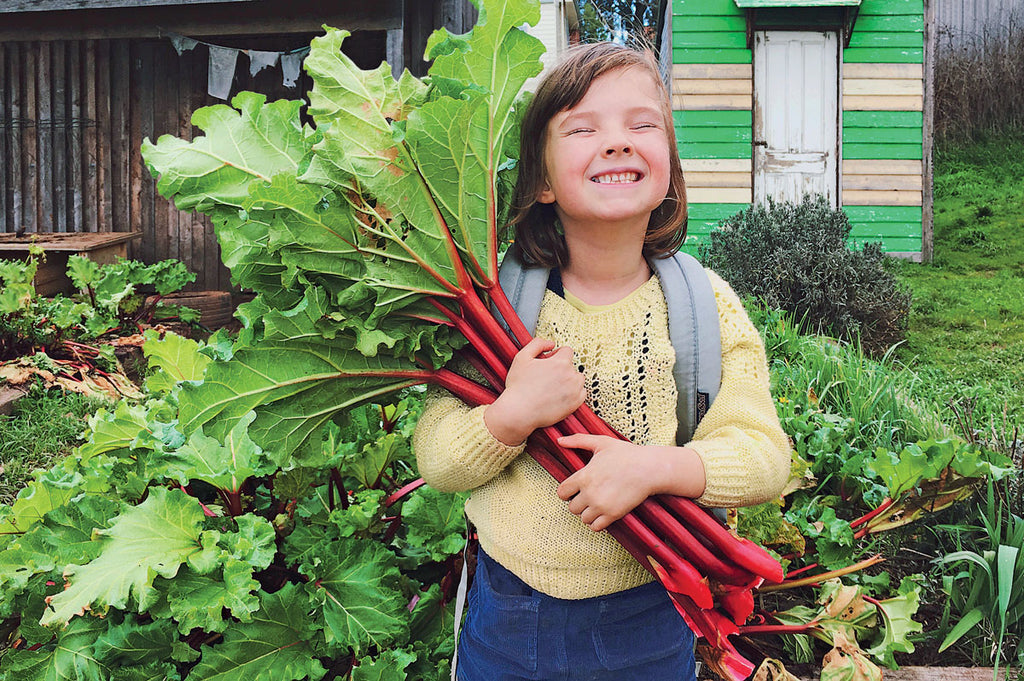 A girl stands in a Stephanie Alexander Kitchen Garden holding stems of rhubarb, smiling for Hello Lunchlady Magazine